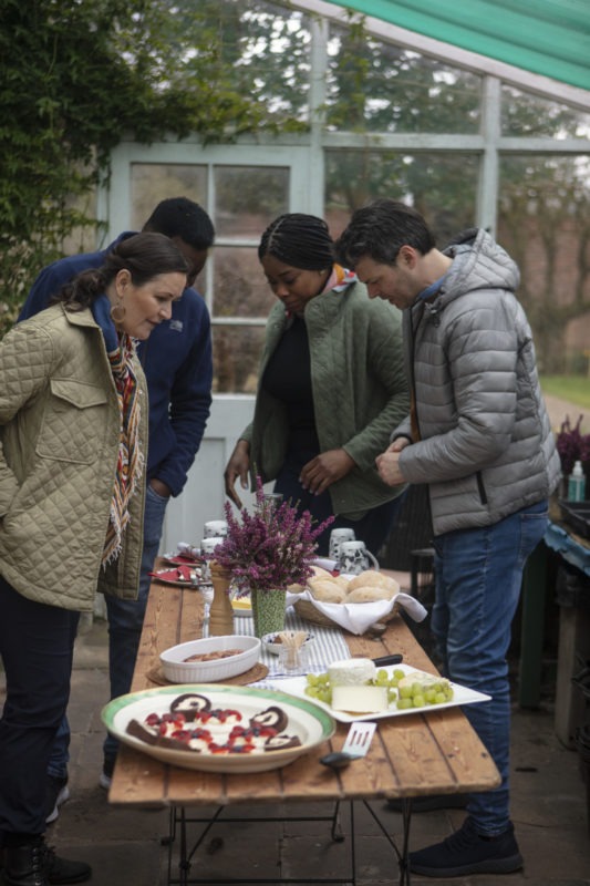 A group of 4 people looking longingly at a table filled with delicious food in a large, Victorian glasshouse. Mini-Sausages, Cheeses and Grapes, Breads.
